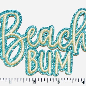 May include: A turquoise glitter embroidered patch with the words "Beach Bum" in a cursive font. The lettering is outlined in a light beige color. The patch is approximately 4 inches wide.