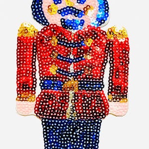 Op de afbeelding: Een kerstversiering in de vorm van een speelgoedsoldaat, met een rode jas, een blauwe broek en een blauwe hoed. De speelgoedsoldaat heeft een roze gezicht, een blauwe snor en gouden accenten. De versiering is ongeveer 7.6 cm hoog.