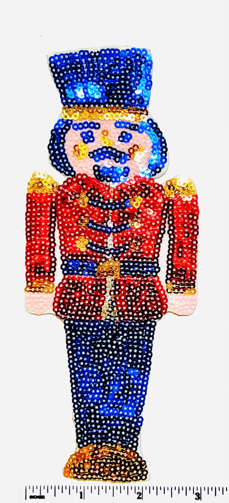 Op de afbeelding: Een kerstversiering in de vorm van een speelgoedsoldaat, met een rode jas, een blauwe broek en een blauwe hoed. De speelgoedsoldaat heeft een roze gezicht, een blauwe snor en gouden accenten. De versiering is ongeveer 7.6 cm hoog.