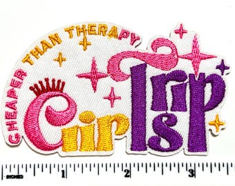 May include: Embroidered patch with the text "Cheaper Than Therapy Girls Trip". The patch features pink, yellow, and purple lettering and star accents on a white background. The patch is approximately 3 inches wide.