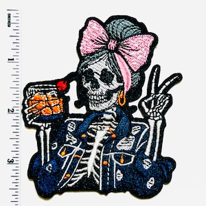 May include: Embroidered patch featuring a skeleton with a pink bow, holding a drink and giving a peace sign. The skeleton wears a blue jacket and gold hoop earrings. The patch is black with white, blue, pink, and orange details.