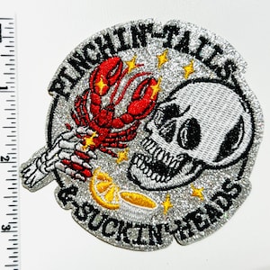 May include: Embroidered patch with the text "PINCHIN' TAILS & SUCKIN' HEADS". The design features a red crawfish held by a skeleton hand, a skull, yellow stars, and a lemon slice. The patch has a silver glitter background.
