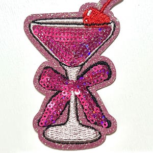 May include: A pink sequined cocktail patch with a red cherry on top. The patch features a bow and a glittery outline. The image includes a ruler with inch measurements.