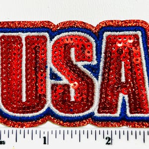 May include: A sequined patch in the shape of the word "USA". The letters are red with white and blue trim, and a glittery red border. The patch is approximately 2 inches wide.