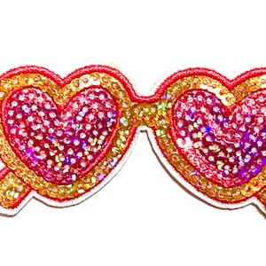 May include: Heart-shaped sunglasses patch with red frames and lenses filled with iridescent sequins. The frames are outlined in gold, and the patch has a white border. A ruler is on the left side.