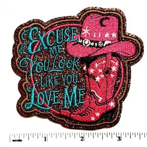 May include: Embroidered patch with a cowboy theme. The patch features a pink cowboy hat and boot with white stars, and the text "EXCUSE ME You Look Like You LOVE ME" in turquoise and pink. The patch has a glittery brown border.