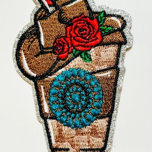 May include: Embroidered patch in the shape of a coffee drink with a red and white striped straw. The brown drink has red roses and a teal floral design. The patch is outlined with silver glitter.