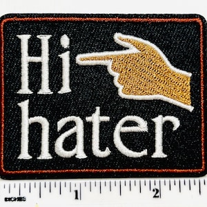 May include: Black rectangular patch with the words "Hi hater" in white, stylized text. A gold-colored hand with a pointing finger is to the right of the word "Hi". The patch has a red border.
