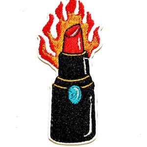 May include: Embroidered patch featuring a black lipstick with a red top, surrounded by flames in shades of red, orange, and yellow. The lipstick has a gold band and a turquoise gem. The patch is approximately 1 inch wide.