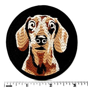 May include: Embroidered patch featuring a dachshund dog's face. The dog has brown and tan fur with large, expressive eyes and a black nose. The patch is circular, with the dog's head centered on a black background. The patch measures approximately 3 inches in diameter.