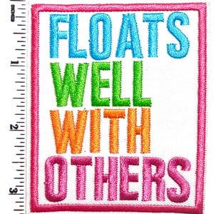 May include: Embroidered patch with the words "FLOATS WELL WITH OTHERS" in blue, green, orange, and pink. The text is on a white background, framed by a pink border. The patch measures approximately 3 inches in height.