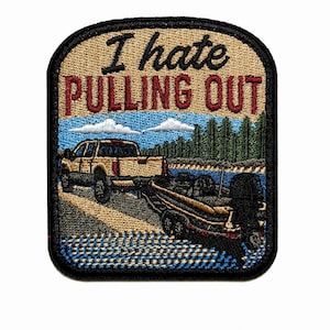 May include: A rectangular embroidered patch with a black border featuring a tan pickup truck towing a boat on a trailer. The text "I hate PULLING OUT" is stitched in black and red. The scene includes a blue sky, green trees, and water.