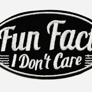 May include: Black oval patch with white embroidered text reading "Fun Fact I Don't Care". The patch has a white border and horizontal lines at the bottom. The patch is approximately 2 inches long.