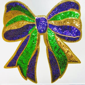May include: A large, decorative bow made of gold, green, and purple sequins. The bow has a gold outline and is designed with alternating stripes of color. The bow is likely used for decoration or embellishment.