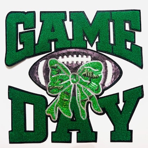 May include: A decorative patch with the words "GAME DAY" in green, outlined in black. A football with a sequined bow is in the center. The football is purple and white. The bow is green and sequined.