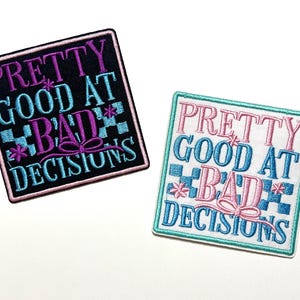 May include: Two embroidered patches with the text "PRETTY GOOD AT BAD DECISIONS". One patch is black with purple and blue accents, the other is white with pink and blue accents. Both have a square shape and a bow design.