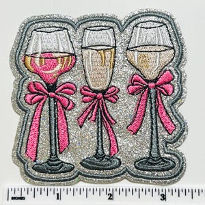 May include: Embroidered patch featuring three champagne glasses with pink bows. The glasses are filled with different colored liquids. The patch has a silver glitter border and is approximately 3 inches wide.