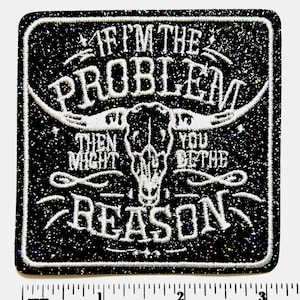 May include: A black, glittery patch with white text and a bull skull design. The text reads "IF I'M THE PROBLEM THEN YOU MIGHT BE THE REASON." The patch is square with rounded corners and a white border. The ruler at the bottom measures up to 3 inches.
