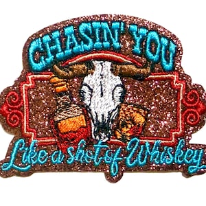 May include: A brown glitter patch with the text "CHASIN' YOU" and "Like a shot of Whiskey" in turquoise. The patch features a steer skull, a whiskey bottle, and a glass of whiskey. The patch measures approximately 2 inches in height.