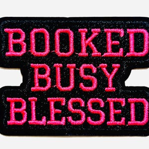 May include: A black embroidered patch with the words "BOOKED BUSY BLESSED" in bright pink thread. The patch is approximately 2 inches in height and 3 inches in width. The patch has a black border.
