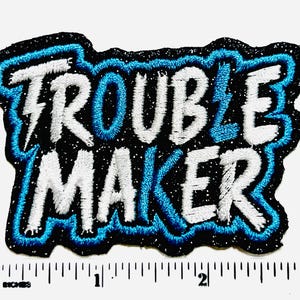 May include: Embroidered patch with the words "TROUBLE MAKER" in white, outlined in blue, on a black background. A blue lightning bolt replaces the letter "L". The patch is approximately 3 inches wide.