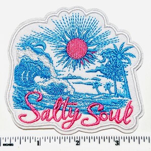 May include: Embroidered patch with a beach scene featuring a pink sun, blue ocean waves, and palm trees. The phrase "Salty Soul" is written in pink script. The patch has a white border.