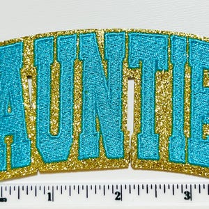May include: Auntie patch with a gold glitter border and turquoise embroidered letters. The patch is arched and measures approximately 3 inches wide. The word "AUNTIE" is displayed in a bold, collegiate-style font.