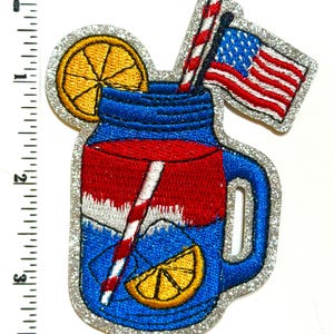 May include: Embroidered patch featuring a patriotic drink design. The patch depicts a blue jar with red and white layers, a straw, and a slice of lemon. An American flag and a silver glitter border complete the design.