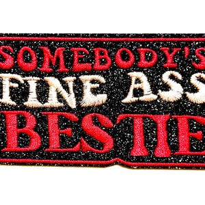 May include: Embroidered patch with the text "SOMEBODY'S FINE ASS BESTIE" in red and off-white thread on a black glitter background. The patch is rectangular with rounded corners and a red border.
