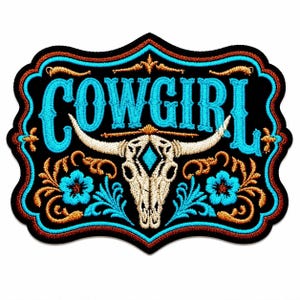 May include: A black embroidered patch featuring the word COWGIRL in bright blue letters. Centered below is a white steer skull with a blue diamond shape on the forehead, flanked by two blue flowers and decorative gold-colored scrollwork.