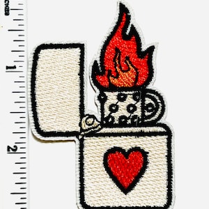 May include: Embroidered patch featuring an open lighter with a red heart and a flame. The lighter is cream-colored with black trim and a red heart. The flame is red and orange. The patch measures approximately 2 inches in height.