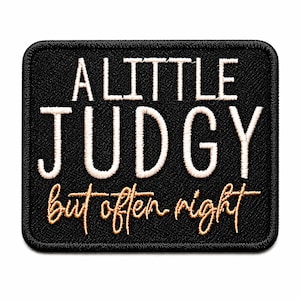 May include: Black rectangular patch with white and gold embroidered text. The text reads "A LITTLE JUDGY but often right". The patch has a black border and is likely an iron-on or sew-on patch.