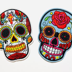 May include: Two embroidered patches in the shape of sugar skulls. One is white with red, yellow, and blue details, and a cross. The other is light blue with a red rose and orange, pink, and yellow details. Both have black eye sockets.