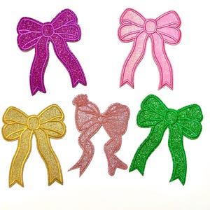 May include: Five glittery bow appliques in various colors: fuchsia, pink, gold, coral, and green. Each bow has a stitched outline and a sparkly texture. These decorative bows are ideal for embellishing crafts, clothing, or accessories.