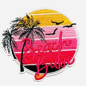 May include: Embroidered patch with a beach theme. The design features a sunset in shades of yellow, pink, and red, with palm trees and the words "Beach Babe" in a vibrant pink script. Includes three flying birds.