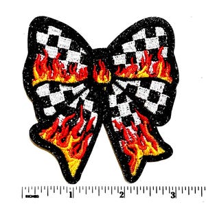 May include: An embroidered bow-shaped patch featuring a black and white checkered pattern overlaid with red and yellow flame designs. The patch has a black glittery border and is positioned above a ruler showing a width of approximately 4 inches.