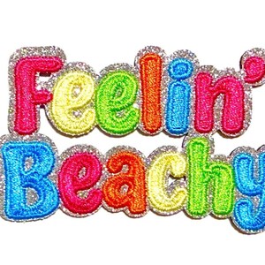 May include: An embroidered patch with the text Feelin' Beachy in multi-colored letters including pink, yellow, green, orange, and blue. The letters are outlined in a silver glitter border. A ruler shows the patch is approximately 2 inches tall.