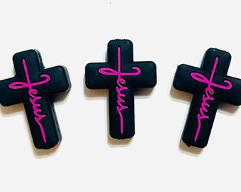 May include: Three black cross charms with the word "Jesus" in pink script. The crosses are arranged on a white surface. The charms are likely for jewelry or crafts.