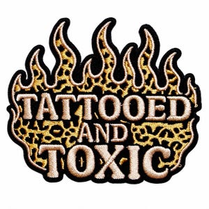 May include: An embroidered patch shaped like flames with a gold and black leopard print pattern. The text TATTOOED AND TOXIC is stitched in white thread across the center of the flame design, outlined with a thick black border for contrast.