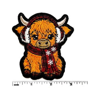 May include: An embroidered patch of a fluffy orange Highland cow wearing red earmuffs and a red scarf with white snowflake patterns. The patch is positioned above a ruler showing a width of approximately 3.5 inches for the design.