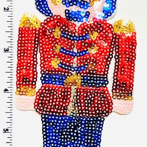 May include: A sequined Christmas ornament in the shape of a toy soldier. The soldier wears a red jacket, blue pants, and a blue hat. Gold accents are on the shoulders and belt. The ornament is approximately 7 inches tall.