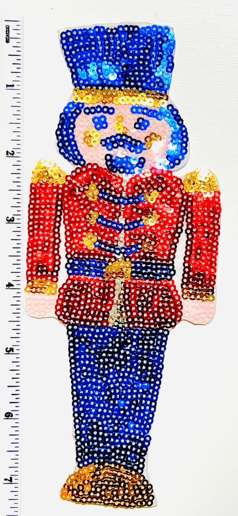 Op de afbeelding: Een kerstversiering in de vorm van een speelgoedsoldaat van pailletten. De soldaat draagt een rode jas, een blauwe broek en een blauwe hoed. Gouden accenten zitten op de schouders en de riem. De versiering is ongeveer 18 cm hoog.