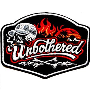 May include: Embroidered patch with a black background and a white border. The patch features a skull wearing a cap and smoking a cigarette, with the word "Unbothered" in white script. Flames and barbed wire complete the design.