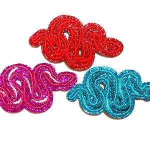 May include: Three glittery, embroidered patches in the shape of a stylized snake. One is red, one is magenta, and the third is turquoise. Each patch has a white embroidered outline and is set against a white background.