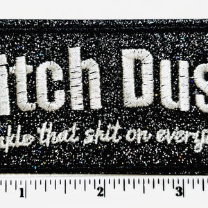 May include: Black rectangular patch with glitter and embroidered white text that reads "Bitch Dust" above "sprinkle that shit on everything". The patch is approximately 4 inches wide.