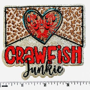 May include: Embroidered patch with a heart-shaped design featuring red crawfish, outlined in teal, on a brown patterned background. The text "CRAWFISH JUNKIE" is in red, outlined in silver glitter.