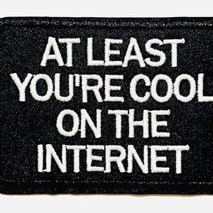 May include: Black embroidered patch with the text "AT LEAST YOU'RE COOL ON THE INTERNET" in white. The patch is rectangular with rounded corners and measures approximately 2 inches in length.