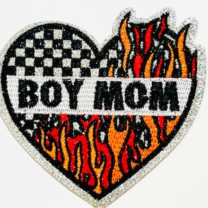 May include: Heart-shaped patch with the words "BOY MOM" in white on a black background. The top half features a black and white checkered pattern, while the bottom half displays flames in red, orange, and yellow. The patch is outlined with silver glitter.