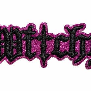 May include: A decorative patch featuring the word Witchy embroidered in black gothic-style lettering. The text is outlined and backed by a vibrant, shimmering magenta glitter material that follows the contour of the letters, creating a bold contrast.