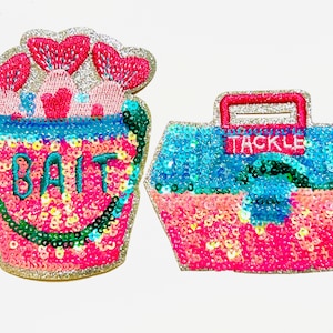 May include: Two sequined patches: a pink bucket with "BAIT" and a tackle box with "TACKLE". The bucket has pink hearts and the tackle box is blue and pink. Both have glittery silver borders.